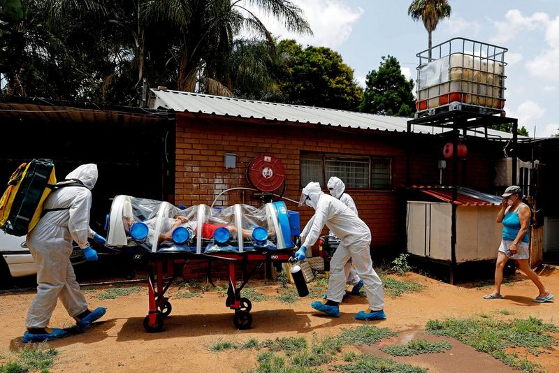 Des ambulanciers poussent un brancard transportant un patient isolé dans une bulle transparente, suivis par une femme portant un masque.