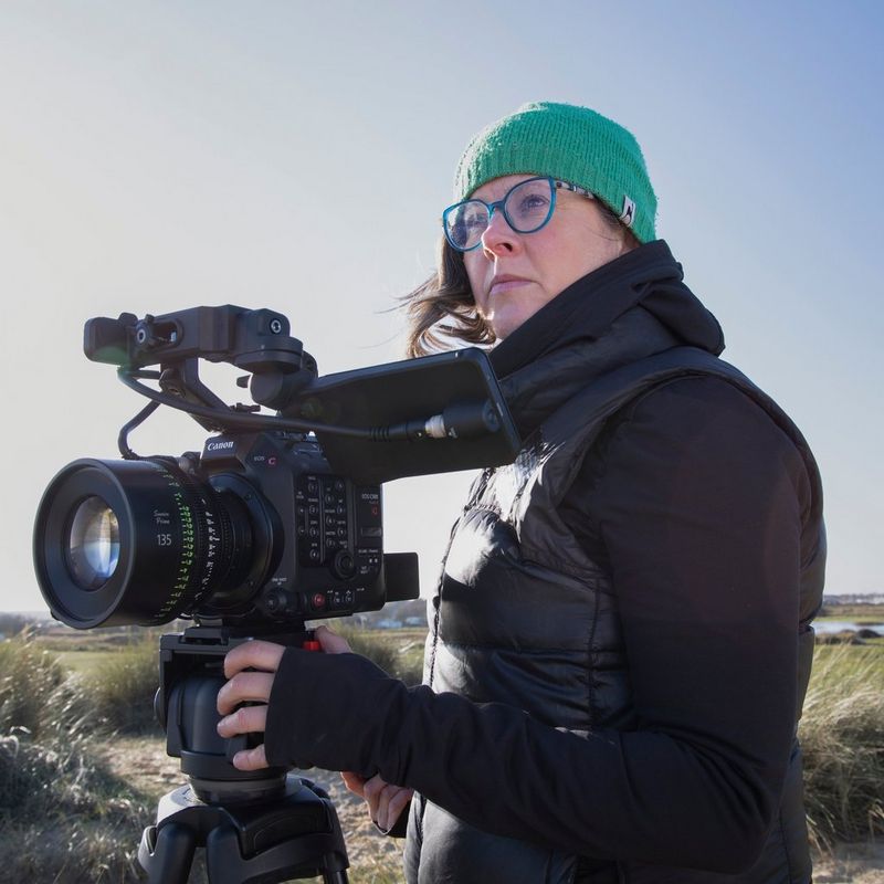  A female cinematographer in a black jacket and green woollen hat standing behind a Canon Cinema EOS camera. 