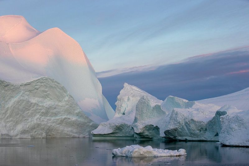 Des icebergs flottant dans une eau calme, le plus grand d'entre eux étant baigné d'une lumière rosâtre émanant du soleil.