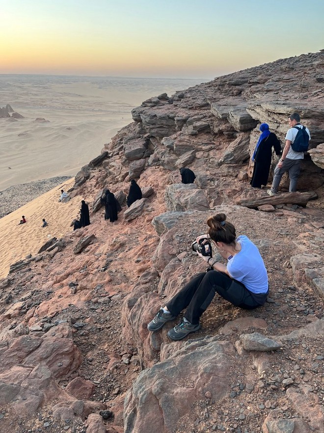 La Canon Ambassador Camilla Ferrari seduta su una sporgenza rocciosa in Sudan, utilizza la sua Canon EOS R5 per scattare fotografie di persone che scendono dalla roccia fino alla spiaggia.