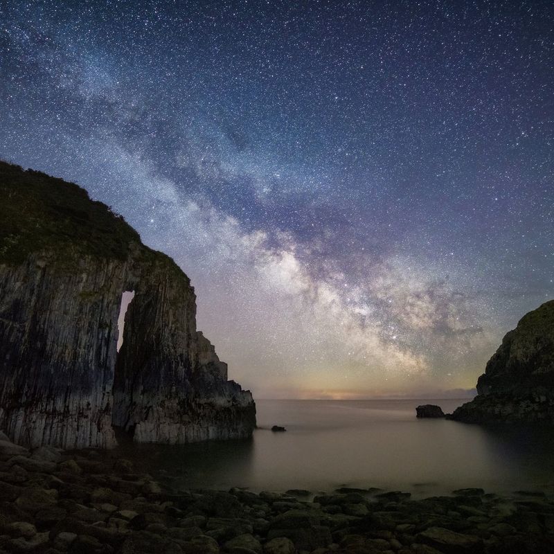 A night sky showing the twinkling Milky Way over the sea, with rocky outcrops on both sides. 