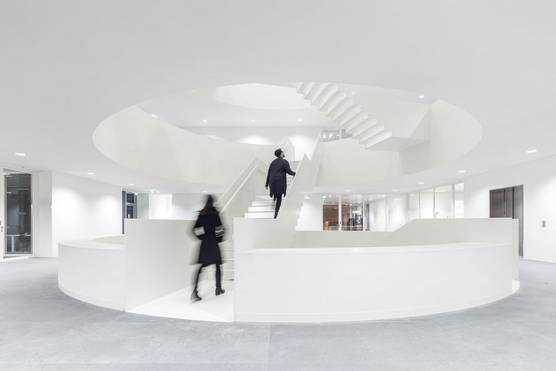 Two figures ascend a circular stairway in the centre of a large white room.
