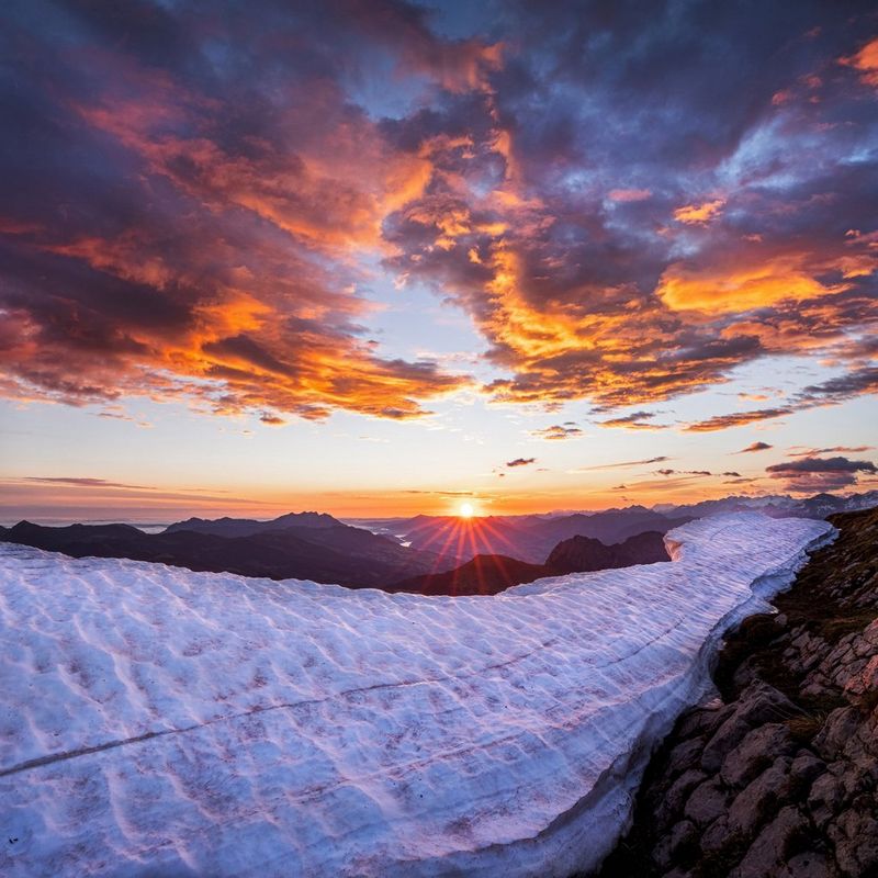 Fluffy clouds and a rocky mountain range, frozen in places, are bathed in an orange glow emanating from the setting sun in the distance. 
