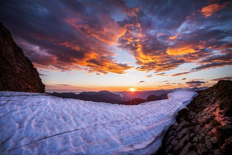 Des nuages cotonneux et une chaîne de montagnes rocheuses, gelée par endroits, sont baignés d'une lueur orange émanant du soleil se couchant au loin.