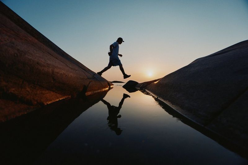 Alors que le soleil se couche à l'horizon, un homme saute d'un gros rocher à un autre. On aperçoit son reflet dans l'eau calme entre les rochers.