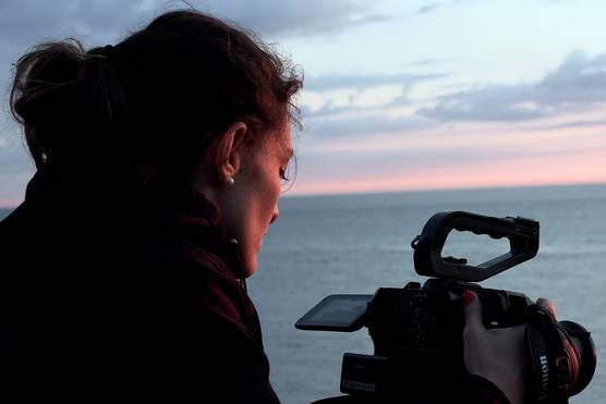 A close up of filmmaker Elisa Iannacone looking at the vari-angle touchscreen of a Canon EOS C70 camera with an expanse of water and pink-hued skyline in front of her.