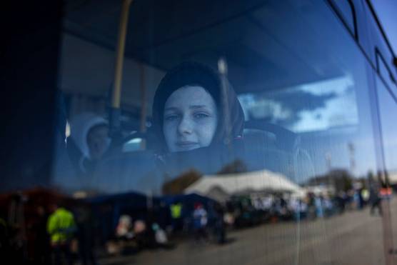 A girl looks out from a stationary bus window. Reflected in the glass are lots of large tents and several people wearing high-vis clothing.