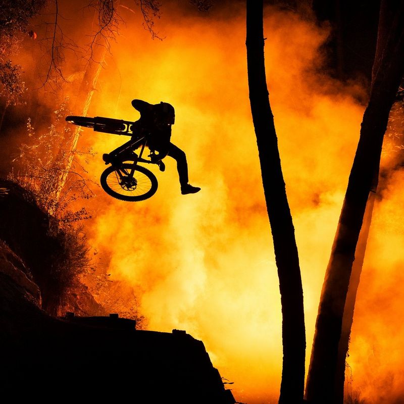 A cyclist is captured in mid air, silhouetted against a smoky orange background, with silhouetted trees and rocks in the foreground.