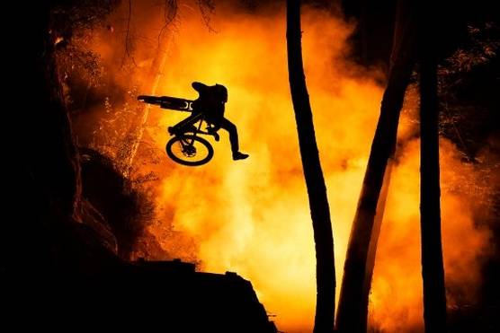 A cyclist is captured in mid air, silhouetted against a smoky orange background, with silhouetted trees and rocks in the foreground.