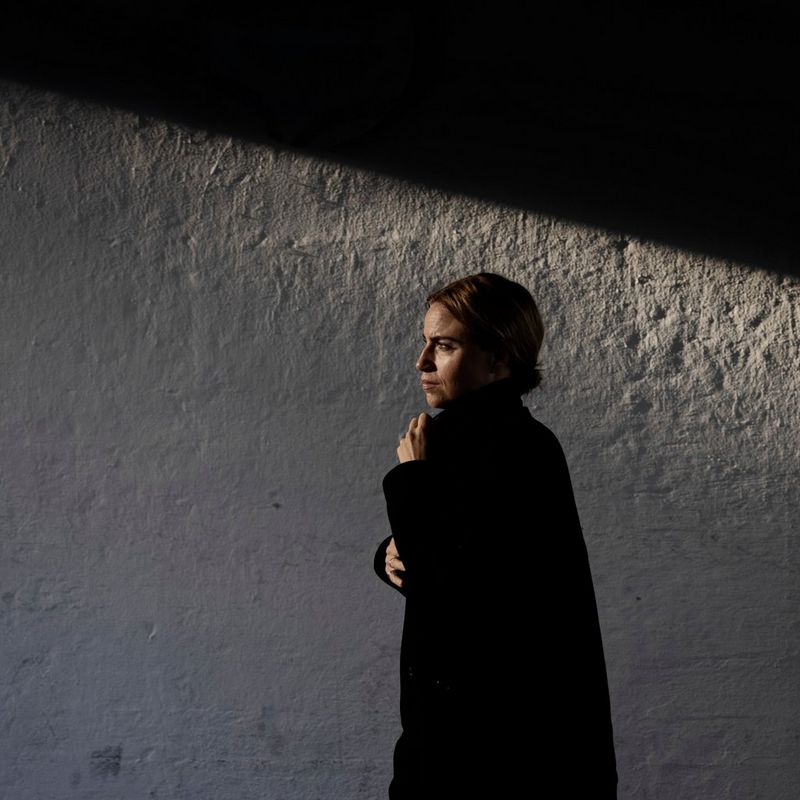 A woman walks along, passing the white painted side of a building. She is holding her coat closed tight around her with both hands.