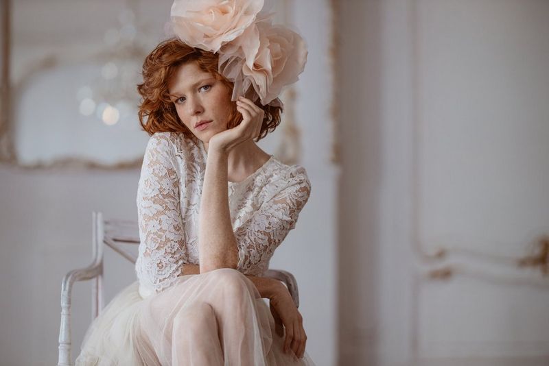 A model sitting posed on a chair and wearing a large peach flower fascinator. 