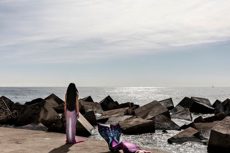 Two women dressed as mermaids gazing out at sea over the rocks. 