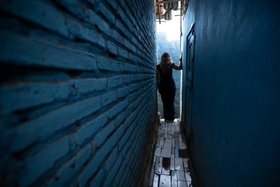 A woman stands at the end of a narrow outdoor passageway, facing away from the camera and leaning against one wall while placing her hand and upper arm against the other.