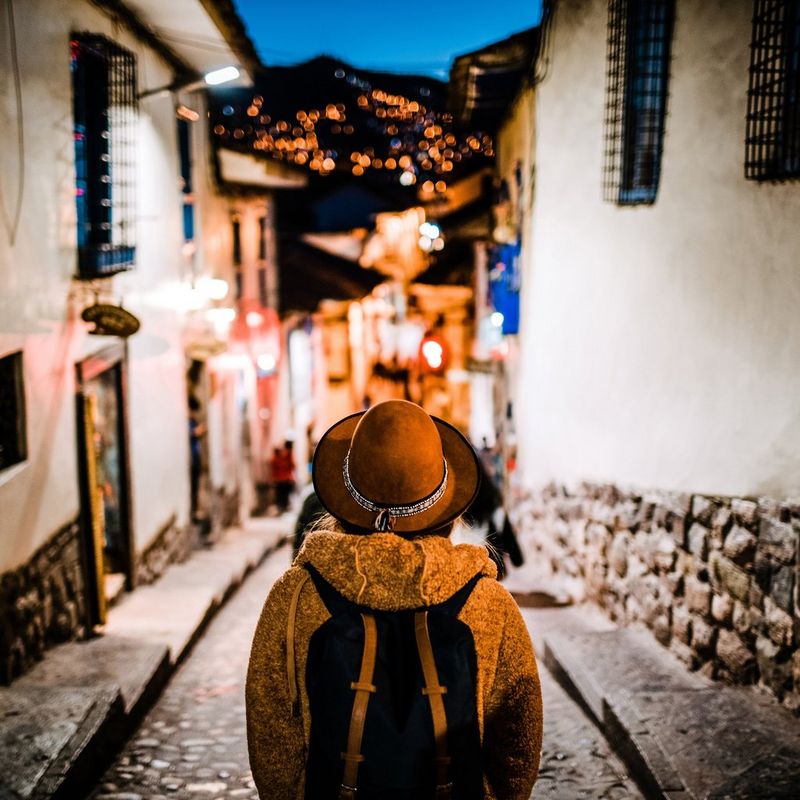 A tourist, wearing a coat, a backpack and a hat, pictured from behind facing an old cobbled street at evening with lights twinkling on a dark hill in the background.