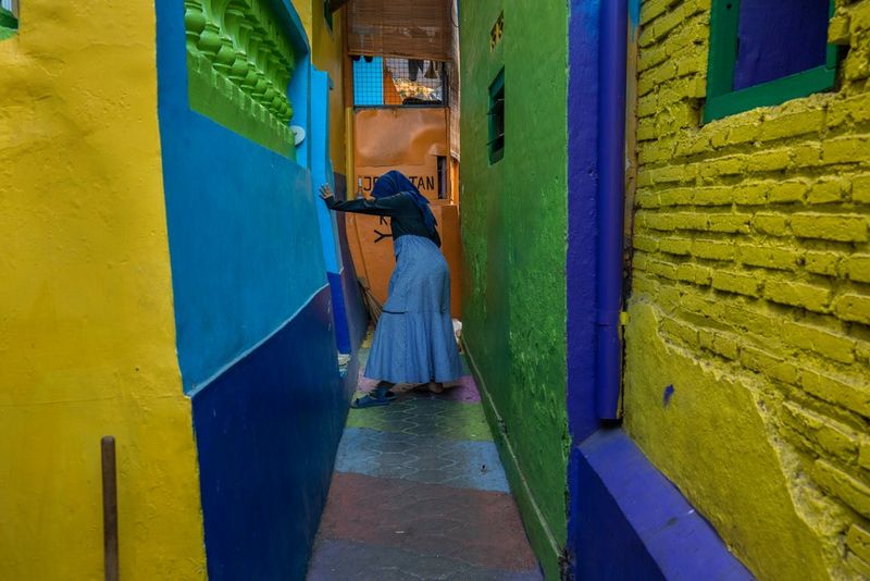 A woman stands at the end of a narrow passageway, facing to the side and away from the camera and leaning forward against one wall, her arm outstretched in front of her. The walls and floor are painted in bright green, blues and yellow.