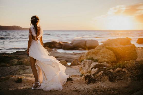 A bride in a long, flowing white dress stands on a rocky coastline, with her back to the camera, as the sun sets over the ocean.