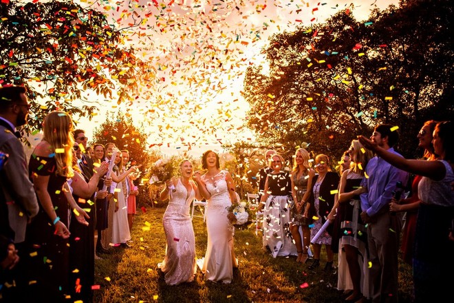 Two brides are showered with confetti by guests in a garden setting in warm late afternoon light.