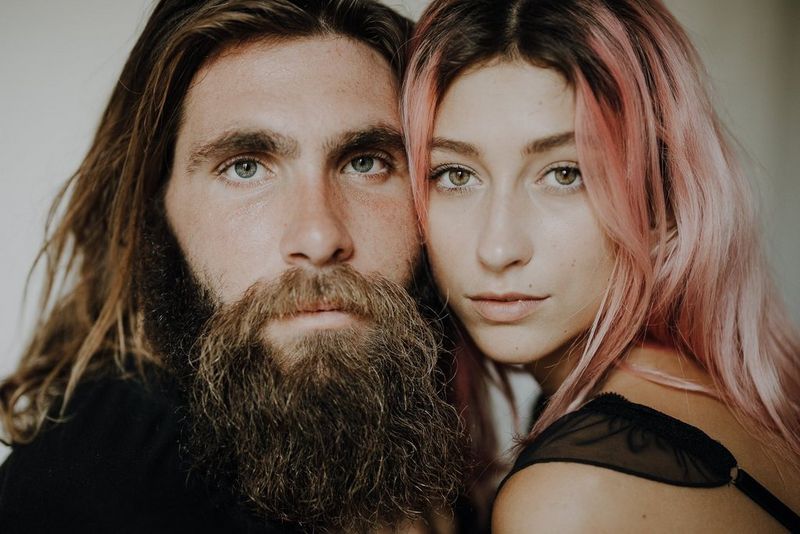 A close-up portrait of a bearded man with long hair and a woman with pink-dyed hair, her face next to his. 