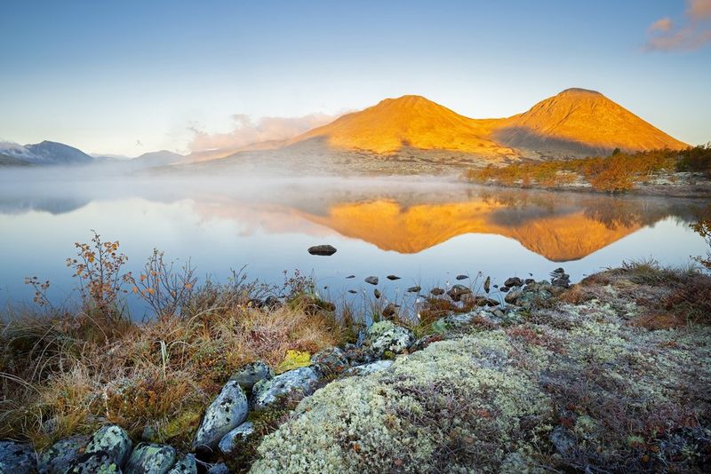 Sand-coloured mountains reflected in a large lake. The rocky shoreline in the foreground is frosty and mist can be seen rising from the still water behind. 