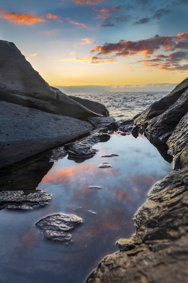 As the sea laps at the shore in the distance, fluffy red clouds reflect in the still water of a rock pool.