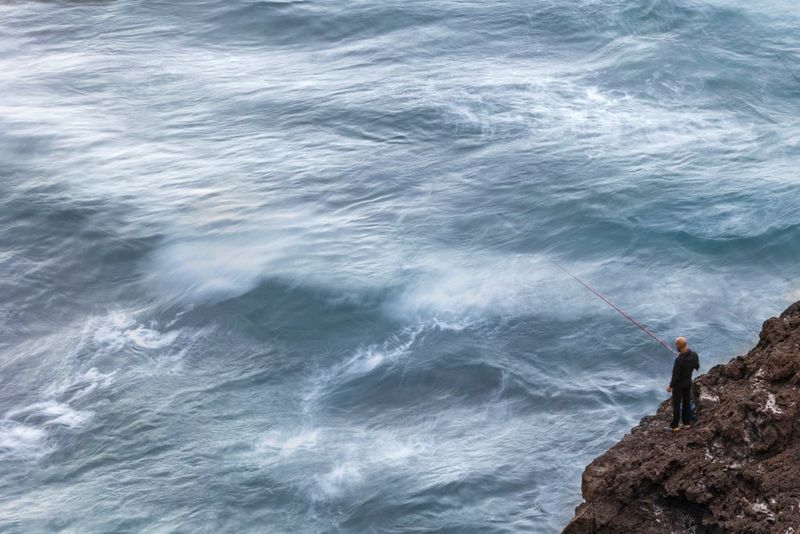 Photographié de haut, un homme seul pêche depuis un affleurement rocheux au-dessus d'une mer agitée.