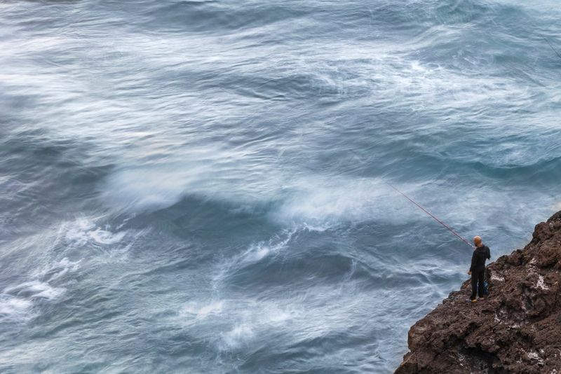 Photographed from above, a lone man fishes from a rocky outcrop above a choppy sea.