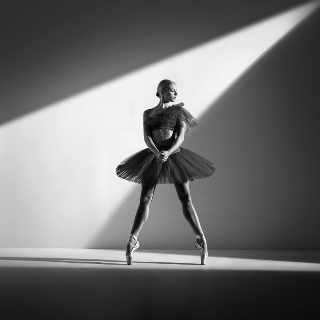 Una ballerina di danza classica sulle punte, con un fascio di luce che le illumina testa e busto mentre il resto del corpo è nell'ombra, è ritratta in uno scatto in bianco e nero di Sascha Hüttenhain, realizzato con Canon EOS R5 e obiettivo Canon RF 24-70mm F2.8 L IS USM.