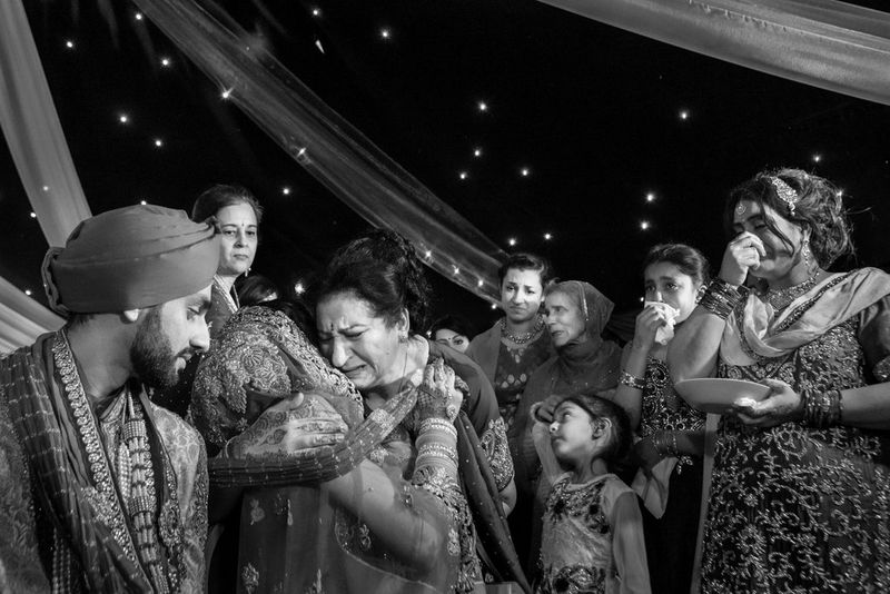 La famiglia della sposa le fa gli auguri di commiato, i parenti la abbracciano e piangono mentre gli altri ospiti osservano la scena in lacrime in questo scatto in bianco e nero realizzato da Sanjay Jogia.