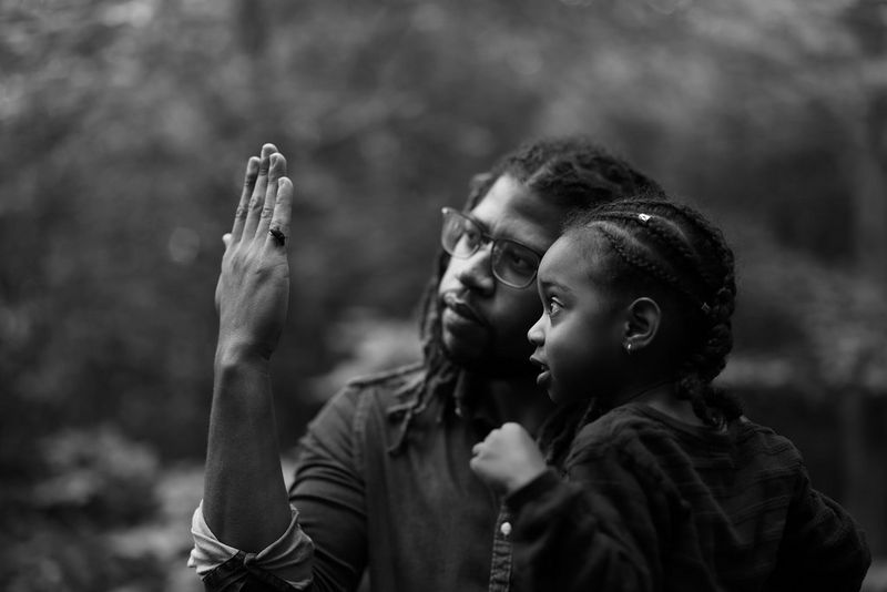 A black and white portrait of a child examining a bug on her father's hand.