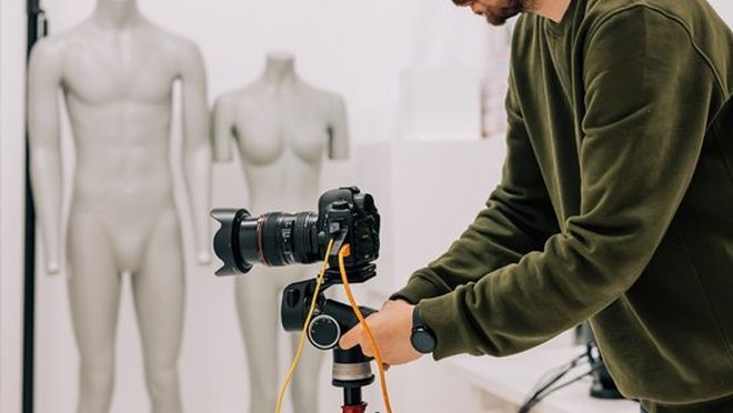A man in a fashion studio adjusts a Canon EOS R5 camera on a tripod, with two mannequins in the background.