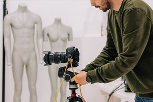A man in a fashion studio adjusts a Canon ֽ_격-5 camera on a tripod, with two mannequins in the background.
