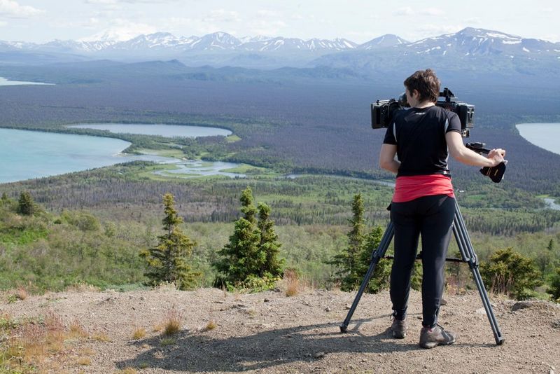 A woman stands on a hill overlooking a valley ringed with mountains. She is operating a camera on a tripod