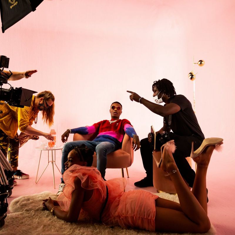 Director Meji Alabi instructs singer Wizkid on the set of his latest music video, while a woman in a short pink dress lies on a rug and another woman in a yellow top adjusts a glass.