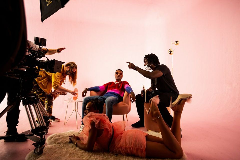 A seated singer on the set of a music video receives instructions from the director. A woman in a short pink dress lies on a rug and another woman in a yellow top adjusts a glass. 
