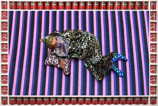 A woman in leopard-spotted clothes with a face covering, lying on a striped pink, black and purple rug. The image's frame consists of red cans with tomatoes on them.