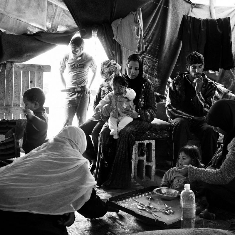 A black and white image of multiple families inside a large tent, with children eating and clothes hanging from lines. 