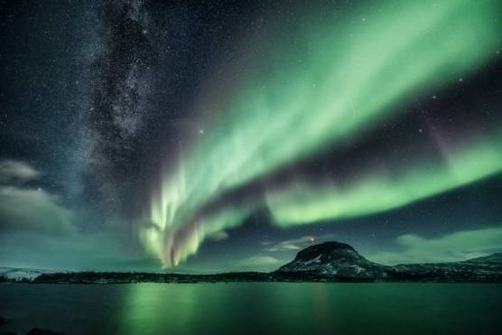 A green aurora lights up the sky above a mountain, the milky way visible next to it.