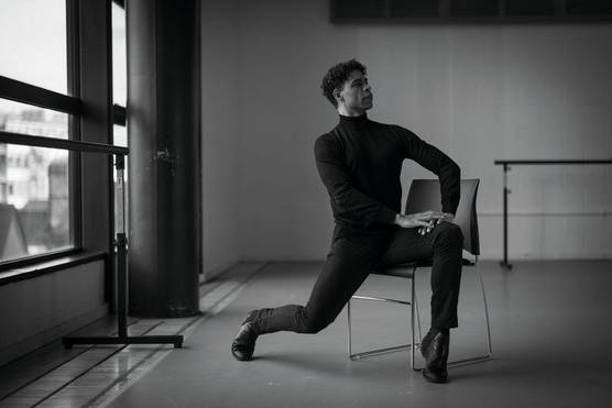A black and white portrait of a man in a black top and trousers posing across a chair in front of a window, leaning forward onto one toe and resting his hands on his leg.