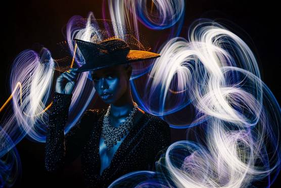 A woman in a low-cut velvet jacket, beaded necklace and large hat tips her hat slightly with her right hand. She is standing mostly in darkness with swirling light trails around and behind her. Taken by Emmanuel Oyeleke with a Canon RF 50mm F1.2L USM lens.
