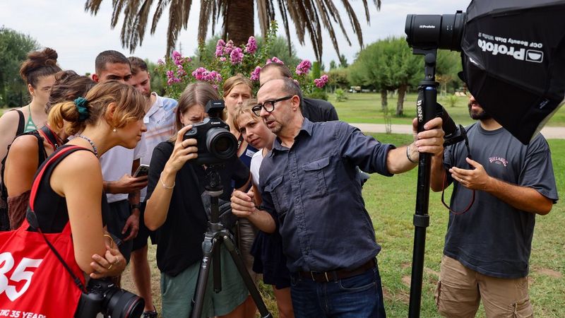 Eine Gruppe versammelt sich um eine Canon Kamera, die auf einem Stativ im Freien montiert ist, und beobachtet aufmerksam, wie der Fotograf Paolo Verzone etwas erklärt, dabei hält er ein Beleuchtungssystem. Im Hintergrund sind Bäume und Blumen.