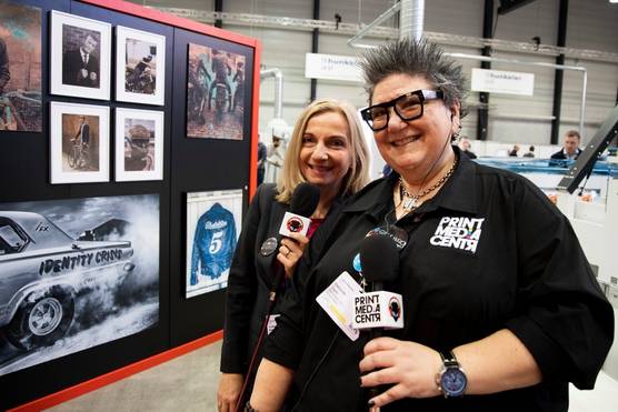 Deborah Corn and Cathy Bittner stand with microphones in front of a photography display stand in a large exhibition hall.