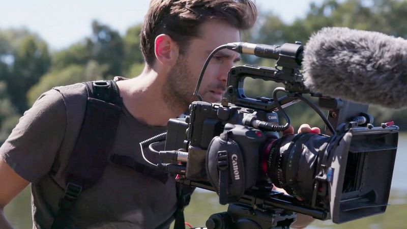 Le directeur de la photographie Ben Sherlock filme avec la caméra Canon EOS C300 Mark III dans un décor rural, avec des buissons et de l'eau en arrière-plan.