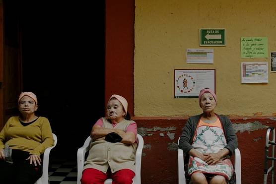 Four older women with beauty facial masks covering their faces sit in a line in front of a yellow and red wall, in this still from a documentary short by filmmaker Irene Baqué.
