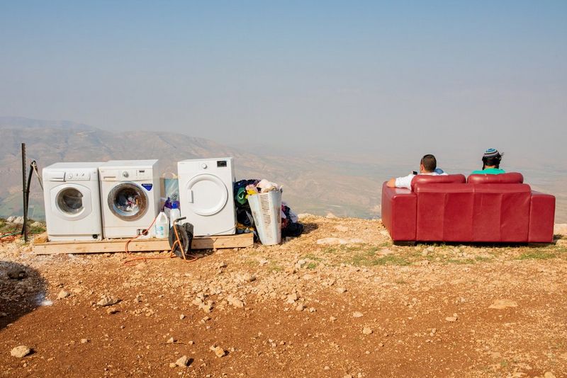Two washing machines and a tumble drier are set up on a pallet on the edge of a dusty mountainside. Next to them is a red sofa, facing away from the camera, with two men sitting on it.