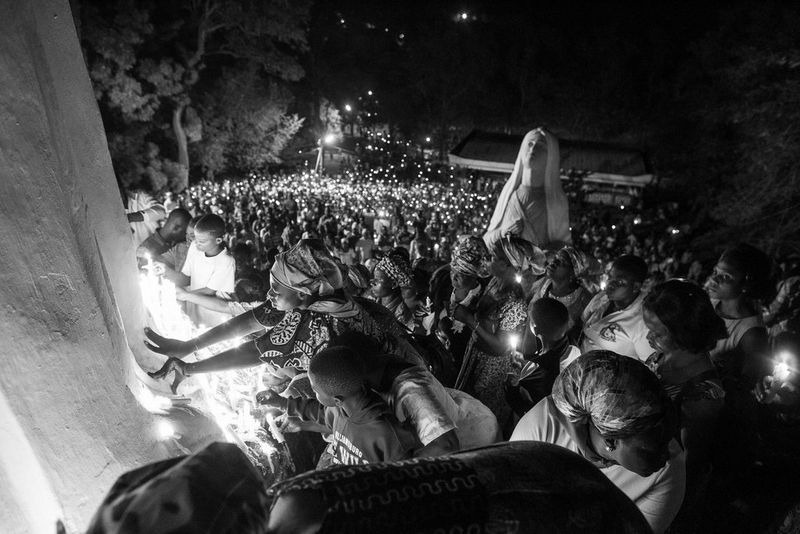 Photo en noir et blanc sur laquelle on peut observer des pèlerins, tenant pour la plupart des bougies, qui se penchent pour toucher les pieds d'une immense statue à la grotte de Notre-Dame de Lourdes, au Ghana.