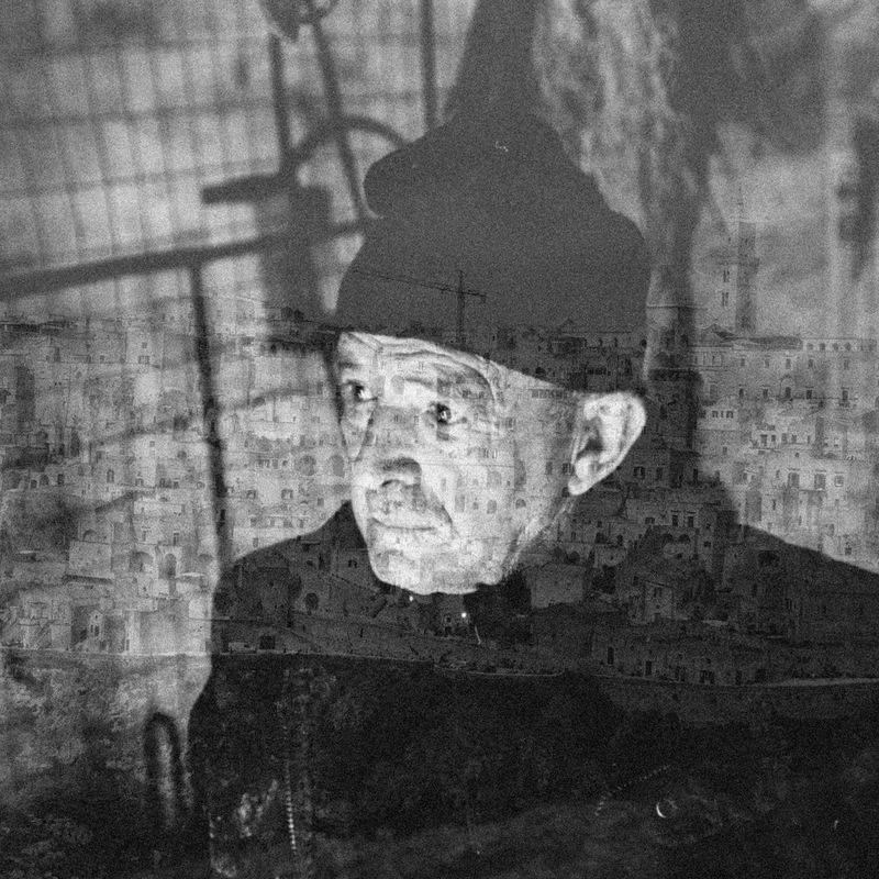 A black and white double exposure of an elderly man in a hat and a cityscape of old stone buildings.
