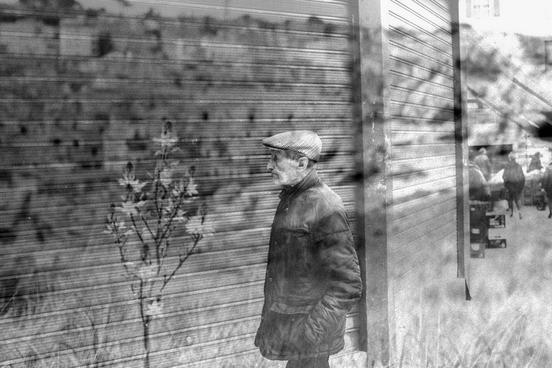 Черно-белая фотография с двойной экспозицией: мужчина в плоской кепке идет вдоль стены + цветение маленького дерева, запечатленного крупным планом.