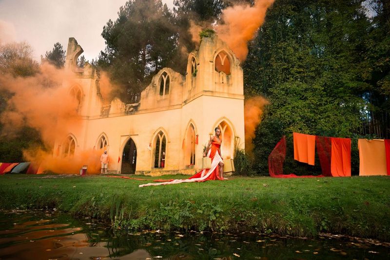 Une femme dans une robe rouge vif marchant devant un château en ruines. Des volutes de fumée orange s'échappent des fenêtres du château.