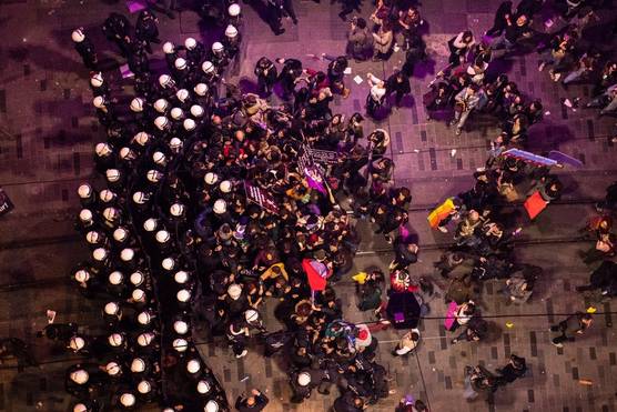 A top-down shot showing several rows of white helmeted police officers blocking the path of a group of protesters carrying purple and pink banners.