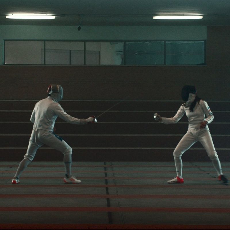 Two fencers sparring in a room lit dimly with fluorescent lighting, one wearing a black mask, the other a silver one.